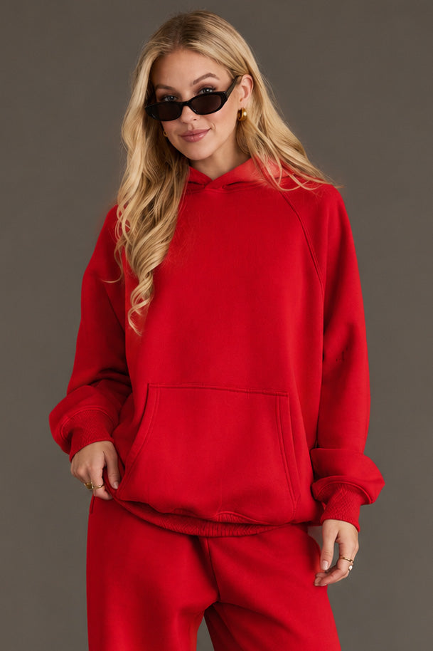 A woman with long blonde hair, wearing black sunglasses and the Red Panache Hooded Sweatshirt from Panache Apparel Collection—featuring a bright red color and soft fleece interior—paired with matching red sweatpants, stands against a plain gray background with her hands in the hoodie pocket.