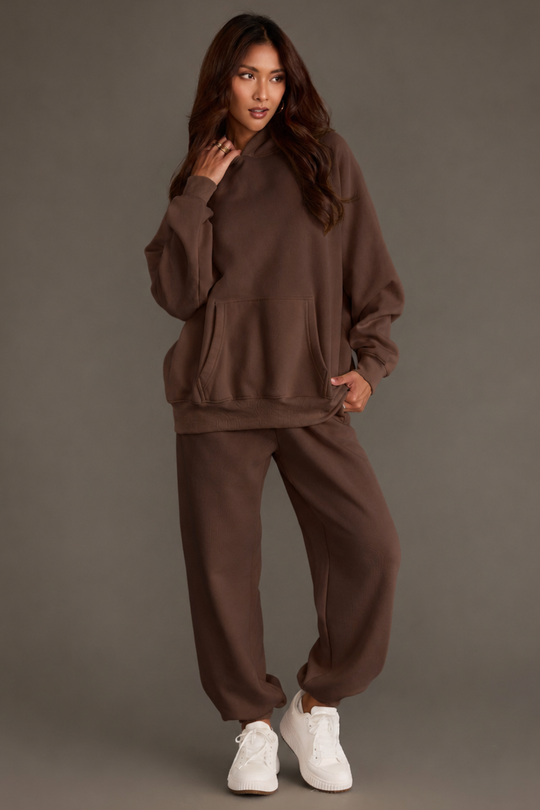 A woman stands against a gray background, wearing a loose Brown Panache Hooded Sweatshirt from the Panache Apparel Collection and matching sweatpants, paired with white sneakers. She has long brown hair and poses with one hand touching her hair and the other in her sweatshirt pocket.