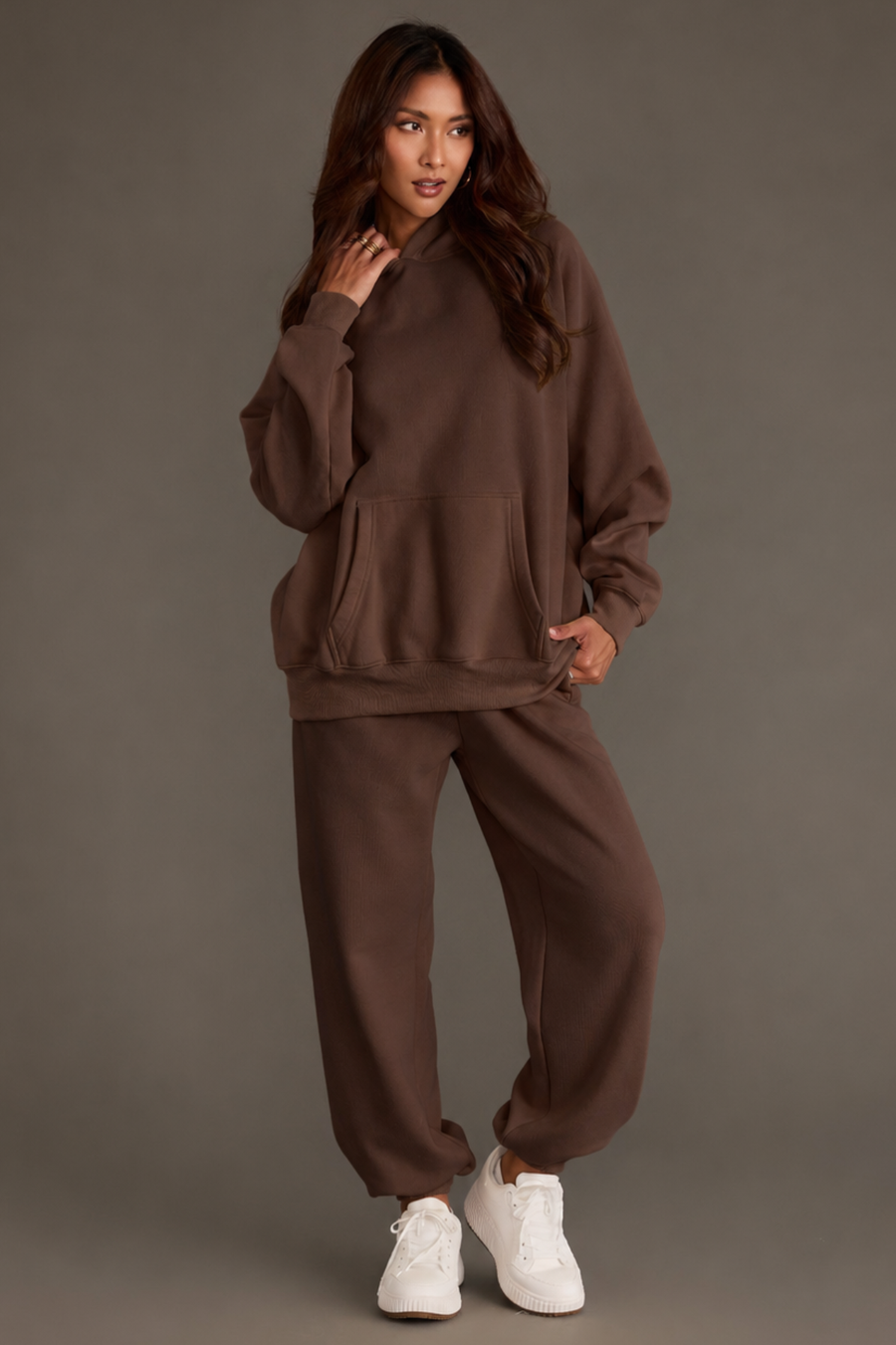 A woman stands against a gray background, wearing a loose Brown Panache Hooded Sweatshirt from the Panache Apparel Collection and matching sweatpants, paired with white sneakers. She has long brown hair and poses with one hand touching her hair and the other in her sweatshirt pocket.
