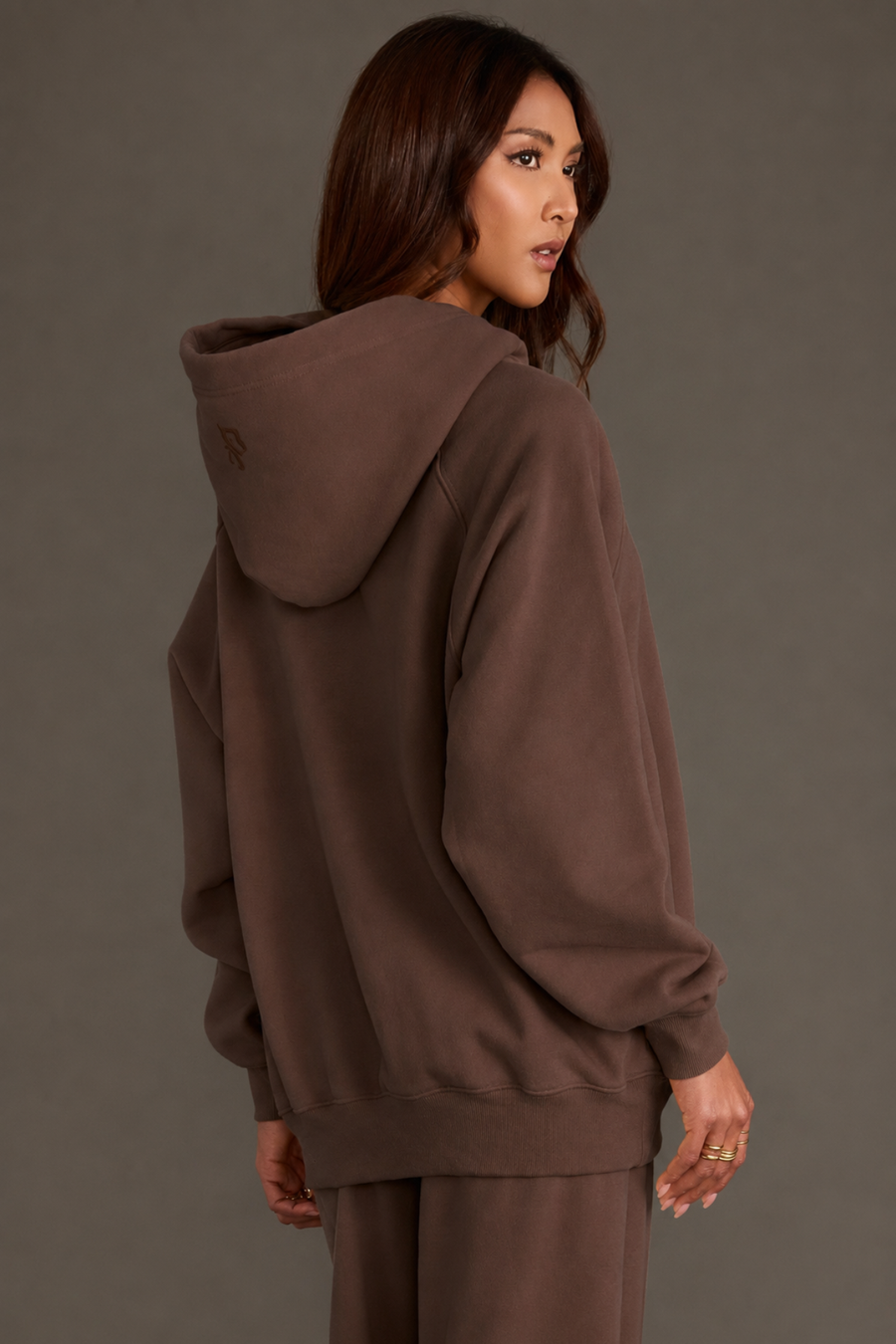 A woman with long brown hair, standing with her back to the camera and head turned to the side against a plain background, is wearing a Brown Panache Hooded Sweatshirt and matching pants from the Panache Apparel Collection.