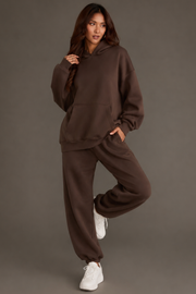 A woman models the Brown Panache Hooded Sweatshirt from the Panache Apparel Collection, paired with matching sweatpants and white sneakers, posing with one leg raised and a hand in her pocket against a plain gray backdrop.