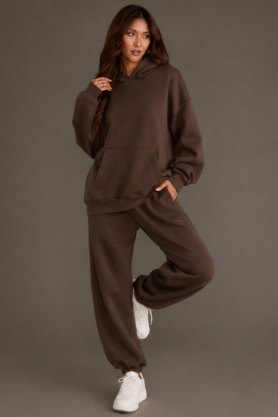 A woman models the Brown Panache Hooded Sweatshirt from the Panache Apparel Collection, paired with matching sweatpants and white sneakers, posing with one leg raised and a hand in her pocket against a plain gray backdrop.
