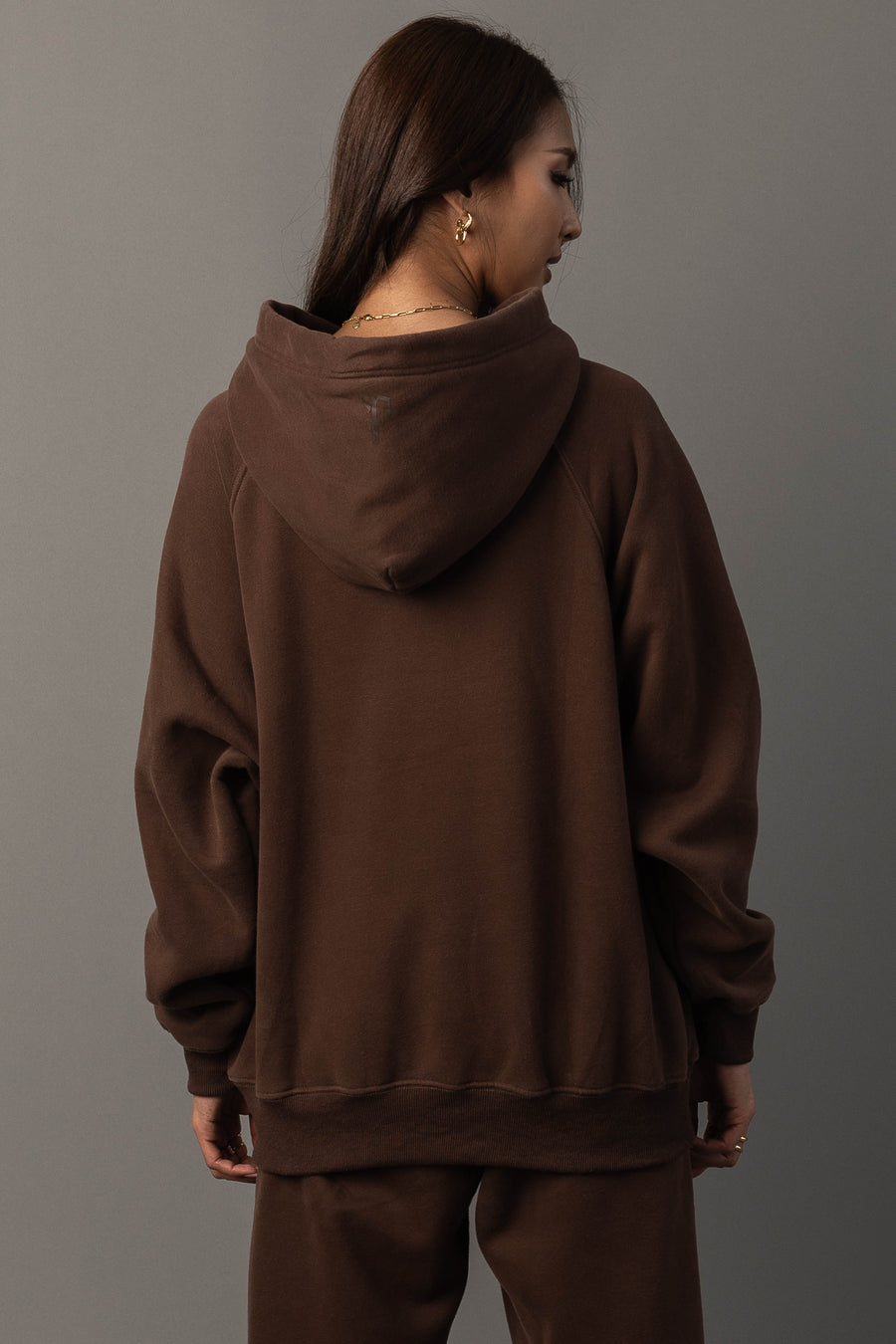 A person with long brown hair is seen from behind, wearing the Brown Panache Hooded Sweatshirt and matching pants from the Panache Apparel Collection, set against a gray background.