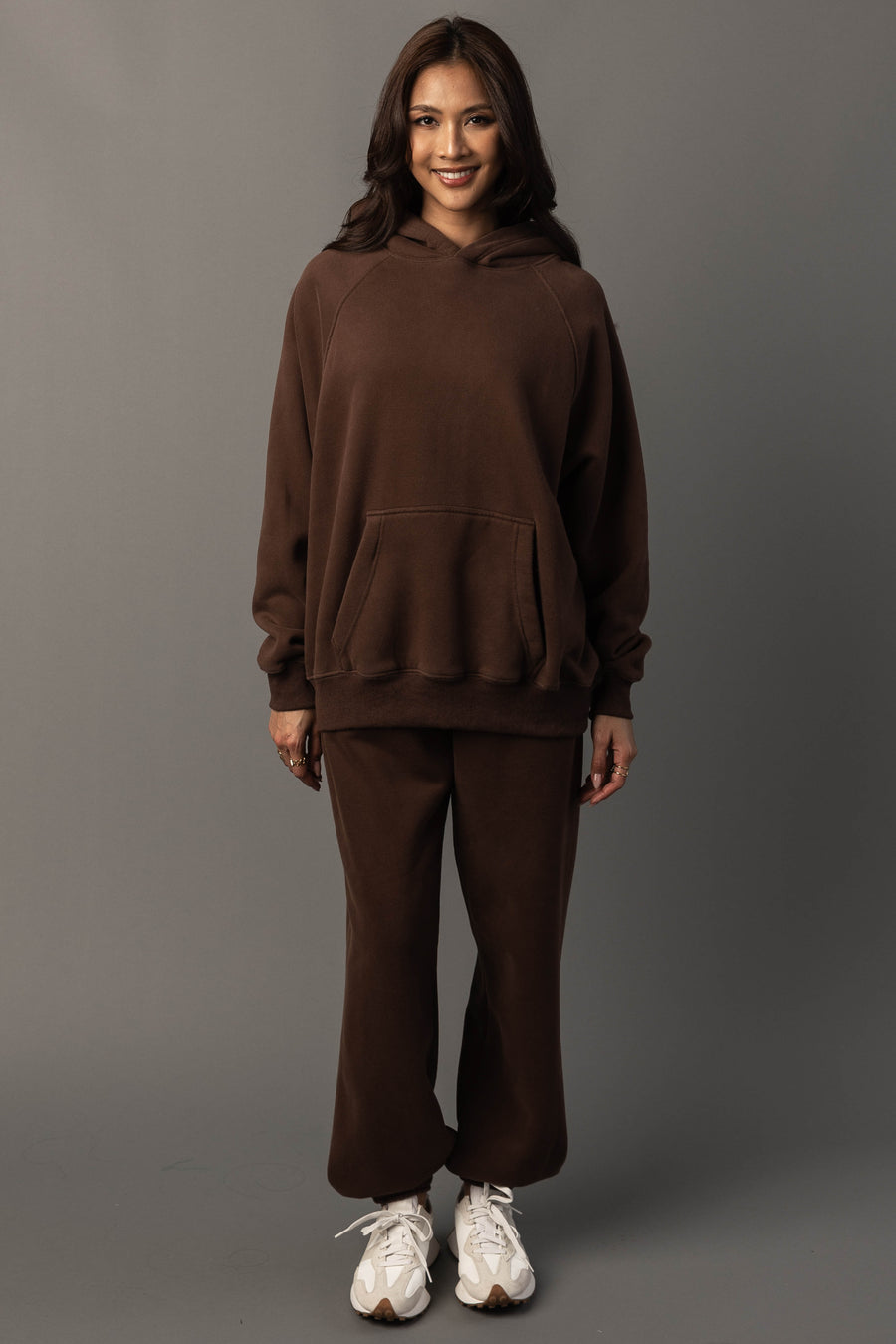 A person stands facing forward, smiling, wearing the Brown Panache Hooded Sweatshirt from Panache Apparel Collection paired with matching sweatpants and white sneakers, set against a plain gray background.