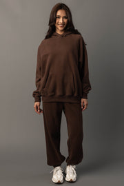 A woman stands smiling against a plain gray background, dressed in the Brown Panache Hooded Sweatshirt and matching sweatpants from the Panache Apparel Collection, paired with white sneakers.