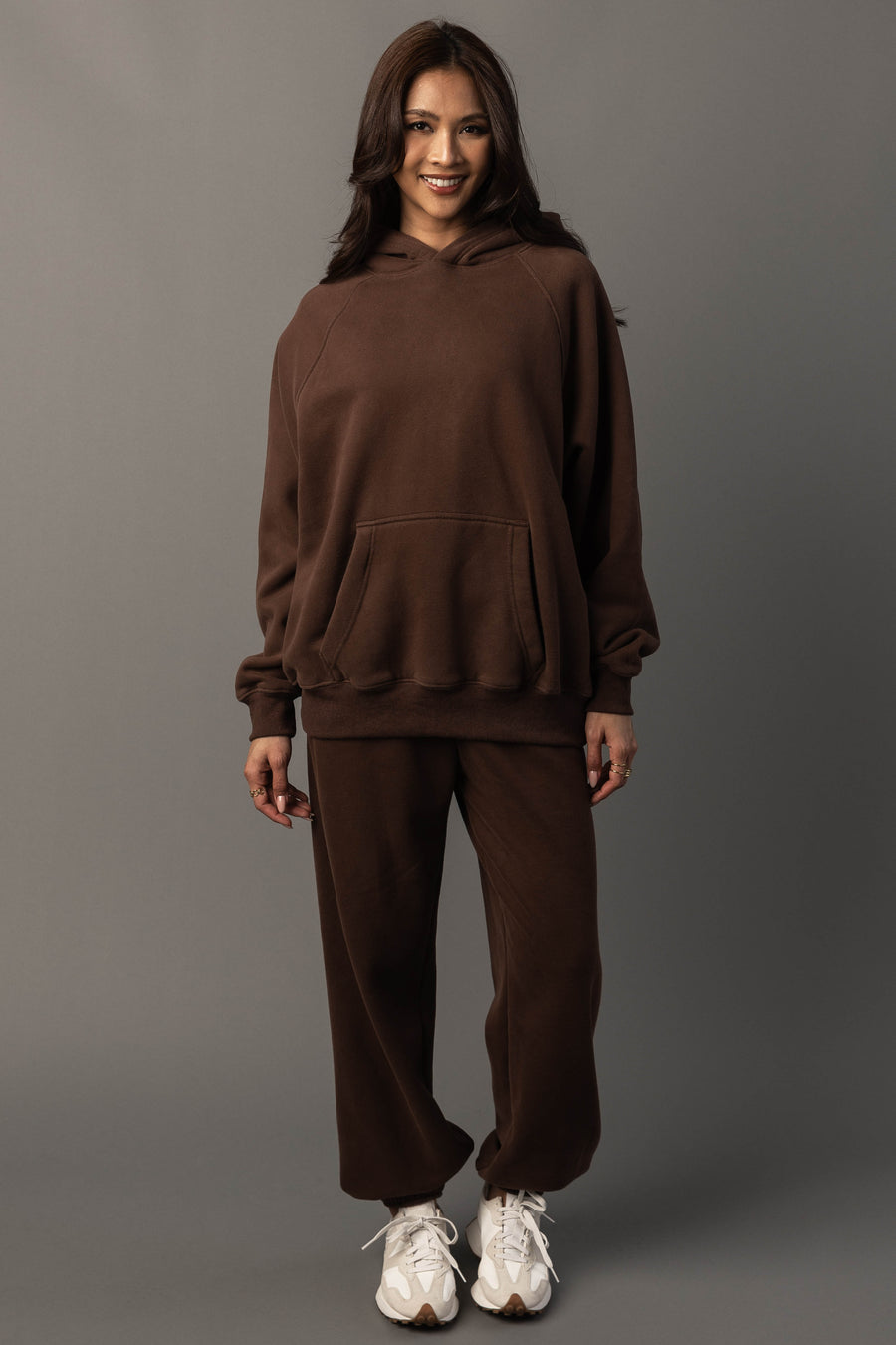 A woman stands smiling against a plain gray background, dressed in the Brown Panache Hooded Sweatshirt and matching sweatpants from the Panache Apparel Collection, paired with white sneakers.