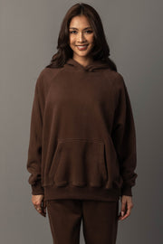 A woman with long brown hair wears the Brown Panache Hooded Sweatshirt and matching pants from the Panache Apparel Collection, standing against a plain gray background and smiling at the camera.