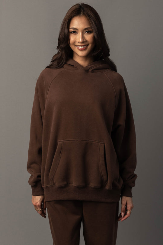 A woman with long brown hair wears the Brown Panache Hooded Sweatshirt and matching pants from the Panache Apparel Collection, standing against a plain gray background and smiling at the camera.