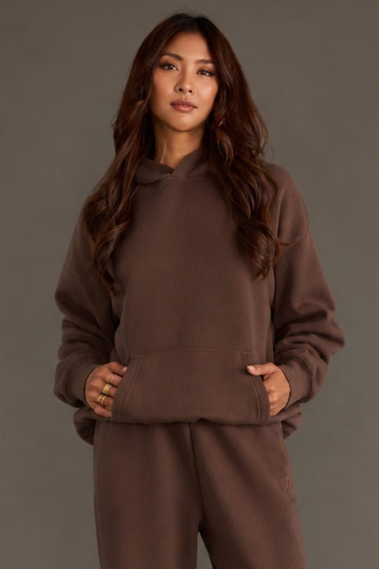 A person with long wavy hair wears a loose-fitting Brown Panache Hooded Sweatshirt and matching pants from the Panache Apparel Collection, standing against a plain gray background with their hands in the hoodie’s pocket.
