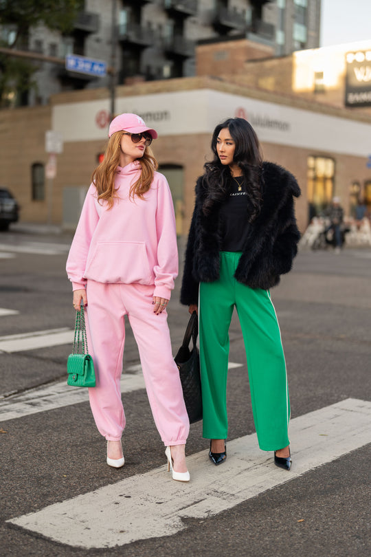 Two women stand on a city crosswalk: one wearing a Light Pink Panache Hooded Sweatshirt with matching sweatpants and a baseball cap, and the other wearing a black fur jacket and green trousers.