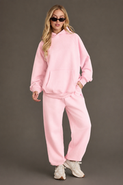 A woman wears the Panache Apparel Collection’s Light Pink Panache Sweatpants with a matching hoodie and white sneakers, standing against a plain background with her hands in her pockets and sunglasses on.
