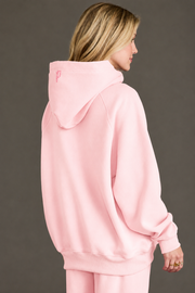 A person with blonde hair, seen from behind, is wearing a loose-fitting Light Pink Panache Hooded Sweatshirt from the Panache Apparel Collection, along with matching pants, against a plain gray background.