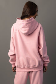 A person with long brown hair, facing away from the camera, is wearing the Panache Apparel Collection’s Light Pink Panache Hooded Sweatshirt and matching pants, standing against a plain gray background.