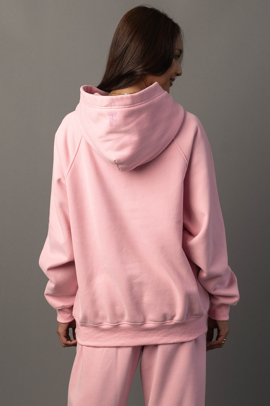 A person with long brown hair, facing away from the camera, is wearing the Panache Apparel Collection’s Light Pink Panache Hooded Sweatshirt and matching pants, standing against a plain gray background.