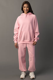 A person stands smiling, wearing the Light Pink Panache Hooded Sweatshirt from the Panache Apparel Collection with matching sweatpants and white sneakers, against a plain gray background.