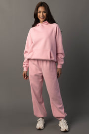 A woman stands smiling against a plain gray background, wearing the Light Pink Panache Hooded Sweatshirt and matching sweatpants from the Panache Apparel Collection, paired with white sneakers.