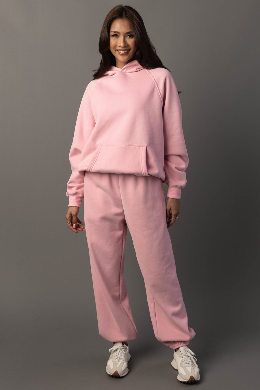 A woman stands smiling against a plain gray background, wearing the Light Pink Panache Hooded Sweatshirt and matching sweatpants from the Panache Apparel Collection, paired with white sneakers.