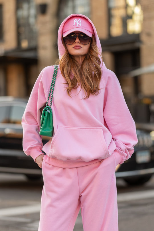 A woman wearing a Light Pink Panache Hooded Sweatshirt, matching sweatpants, a pink baseball cap, and oversized sunglasses, carrying a small green shoulder bag.