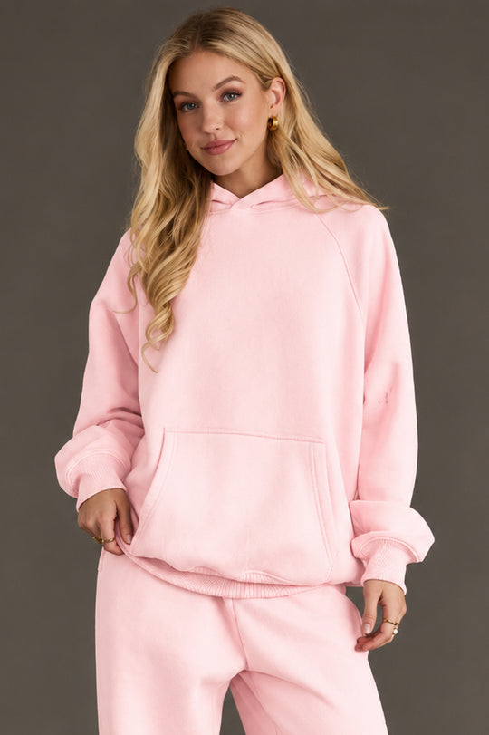 A woman with long blonde hair is wearing the Light Pink Panache Hooded Sweatshirt and matching sweatpants from the Panache Apparel Collection, standing against a plain gray background with her hands in the front pocket of the hoodie.