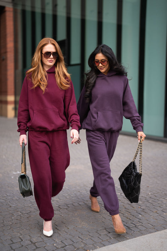 Two women walk side by side wearing matching Plum Panache Hooded Sweatshirts and sweatpants, carrying handbags.