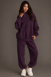 A woman models the Plum Panache Hooded Sweatshirt and matching sweatpants from the Panache Apparel Collection, standing with one hand near her face and gazing to the side. She has long hair, wears white sneakers, and is posed against a plain background.