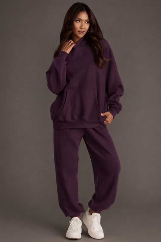 A woman models the Plum Panache Hooded Sweatshirt and matching sweatpants from the Panache Apparel Collection, standing with one hand near her face and gazing to the side. She has long hair, wears white sneakers, and is posed against a plain background.