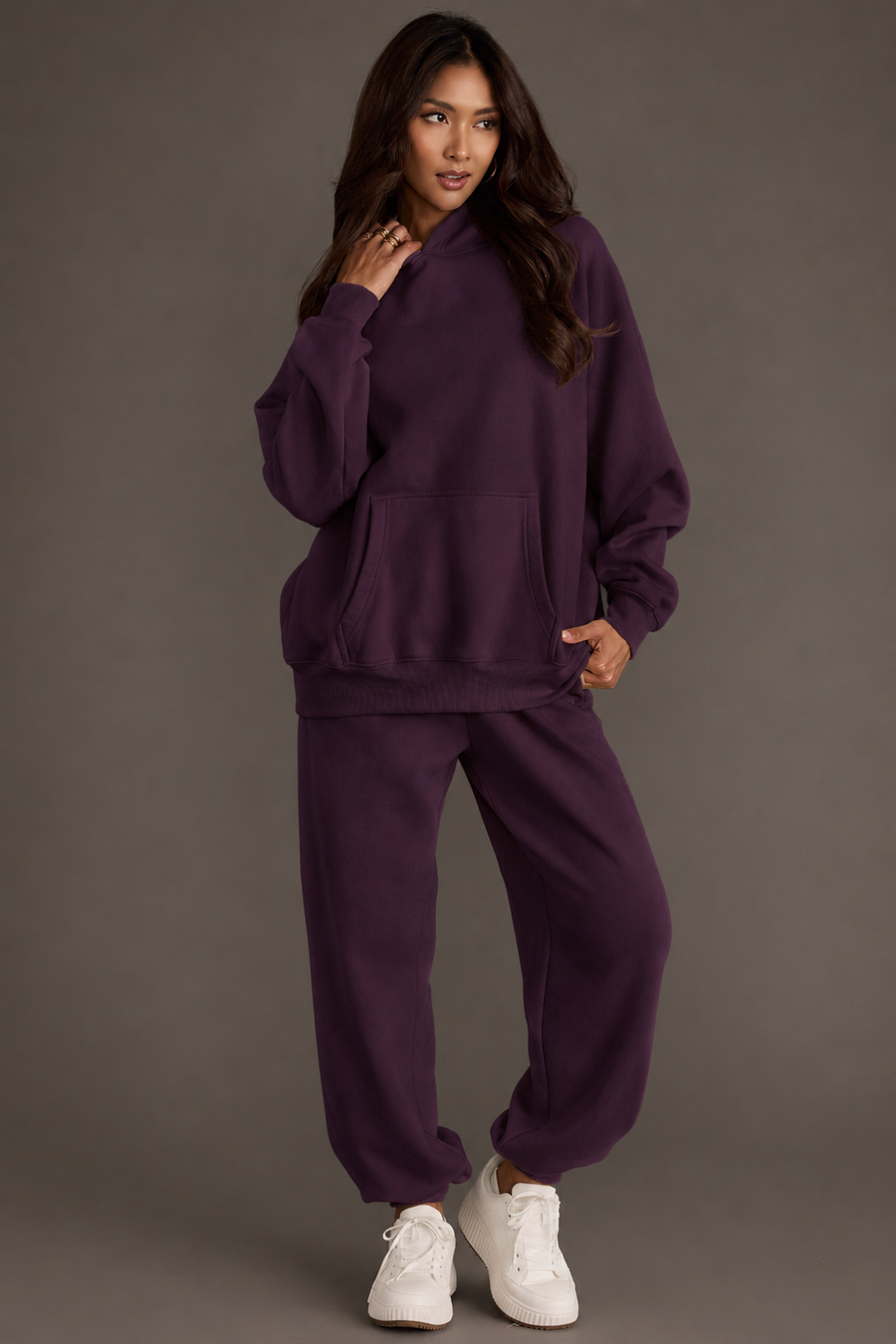 A woman models the Plum Panache Hooded Sweatshirt and matching sweatpants from the Panache Apparel Collection, standing with one hand near her face and gazing to the side. She has long hair, wears white sneakers, and is posed against a plain background.