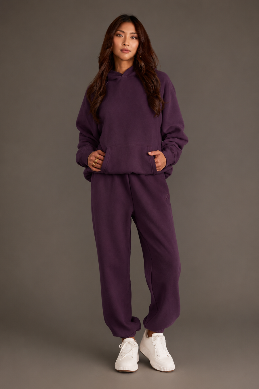 A woman stands against a plain background, wearing the Plum Panache Hooded Sweatshirt from the Panache Apparel Collection paired with matching sweatpants and white sneakers. Her hands are in her hoodie pocket, and she has long, wavy brown hair.