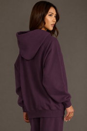 A woman with long brown hair, standing against a plain gray background and looking over her left shoulder, wears the Panache Apparel Collection Plum Panache Hooded Sweatshirt with matching pants.