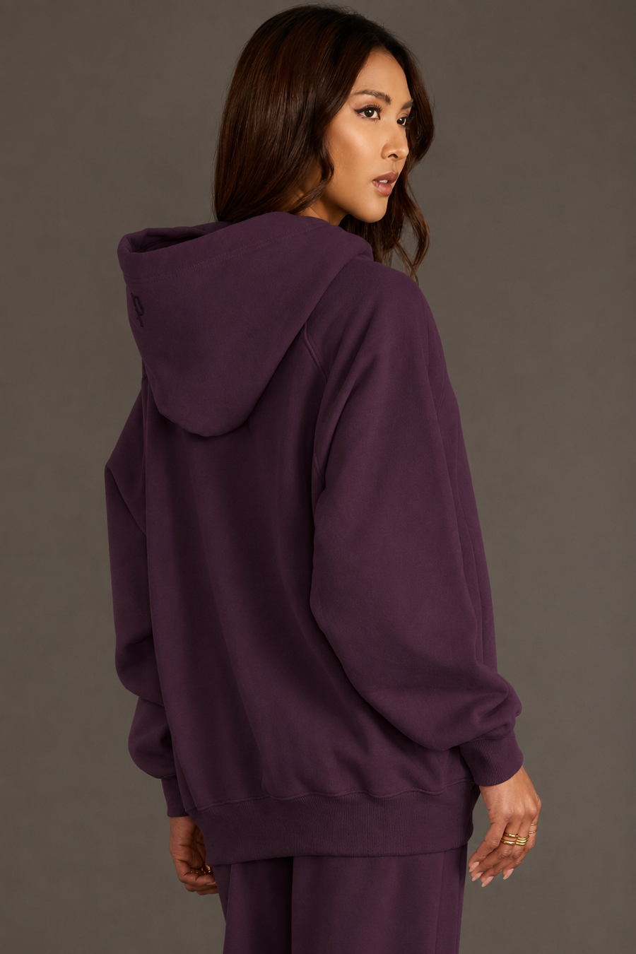 A woman with long brown hair, standing against a plain gray background and looking over her left shoulder, wears the Panache Apparel Collection Plum Panache Hooded Sweatshirt with matching pants.