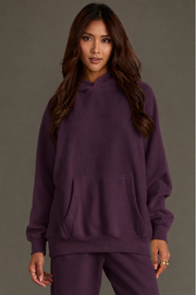 A woman with long brown hair wears the loose-fitting Plum Panache Hooded Sweatshirt from the Panache Apparel Collection, paired with matching pants, as she stands against a plain gray background.