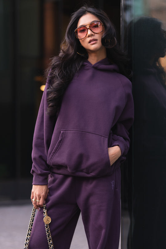 A woman wears a Plum Panache Hooded Sweatshirt and matching sweatpants, with one hand in her pocket and holding a chain-strap handbag in the other.