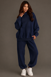 A woman poses casually against a gray background, wearing the Dark Navy Panache Hooded Sweatshirt from the Panache Apparel Collection. The cozy fleece-lined sweatshirt is paired with matching sweatpants and white sneakers as she touches her hair and gazes slightly to the side.