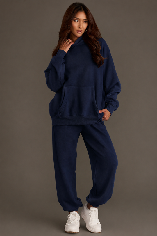 A woman poses casually against a gray background, wearing the Dark Navy Panache Hooded Sweatshirt from the Panache Apparel Collection. The cozy fleece-lined sweatshirt is paired with matching sweatpants and white sneakers as she touches her hair and gazes slightly to the side.