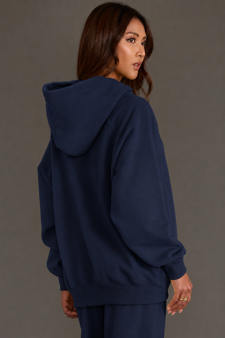 A woman with long brown hair models the Panache Apparel Collection’s Dark Navy Panache Hooded Sweatshirt, which features a cropped relaxed fit and matching pants. She is facing away and looking over her shoulder in front of a plain gray background.