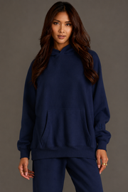 A woman with long brown hair wears the Dark Navy Panache Hooded Sweatshirt from the Panache Apparel Collection, featuring a cozy fleece interior and paired with matching sweatpants. She stands against a plain gray background, facing forward with a neutral expression and her hands relaxed at her sides.