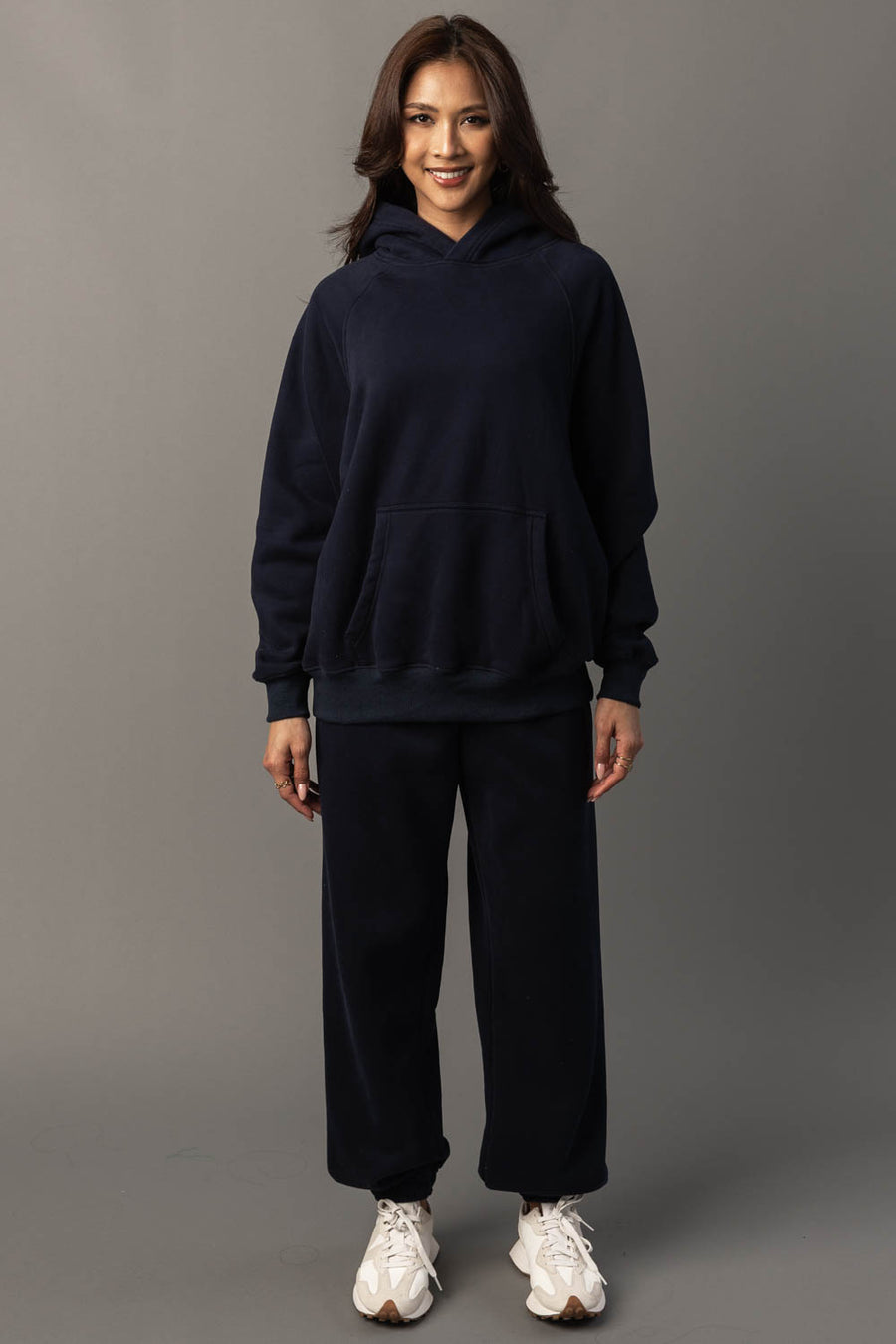 A person stands smiling, wearing the Dark Navy Panache Hooded Sweatshirt from Panache Apparel Collection and matching sweatpants with white sneakers, against a plain gray background.