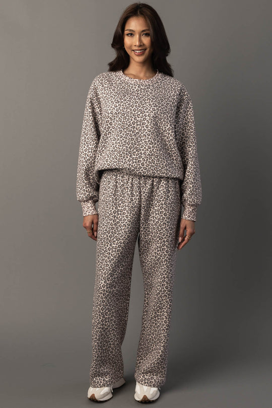 A woman stands smiling against a plain gray background, wearing the Panache Apparel Collection Cream & Brown Leopard Sweatshirt paired with matching pants and white sneakers.