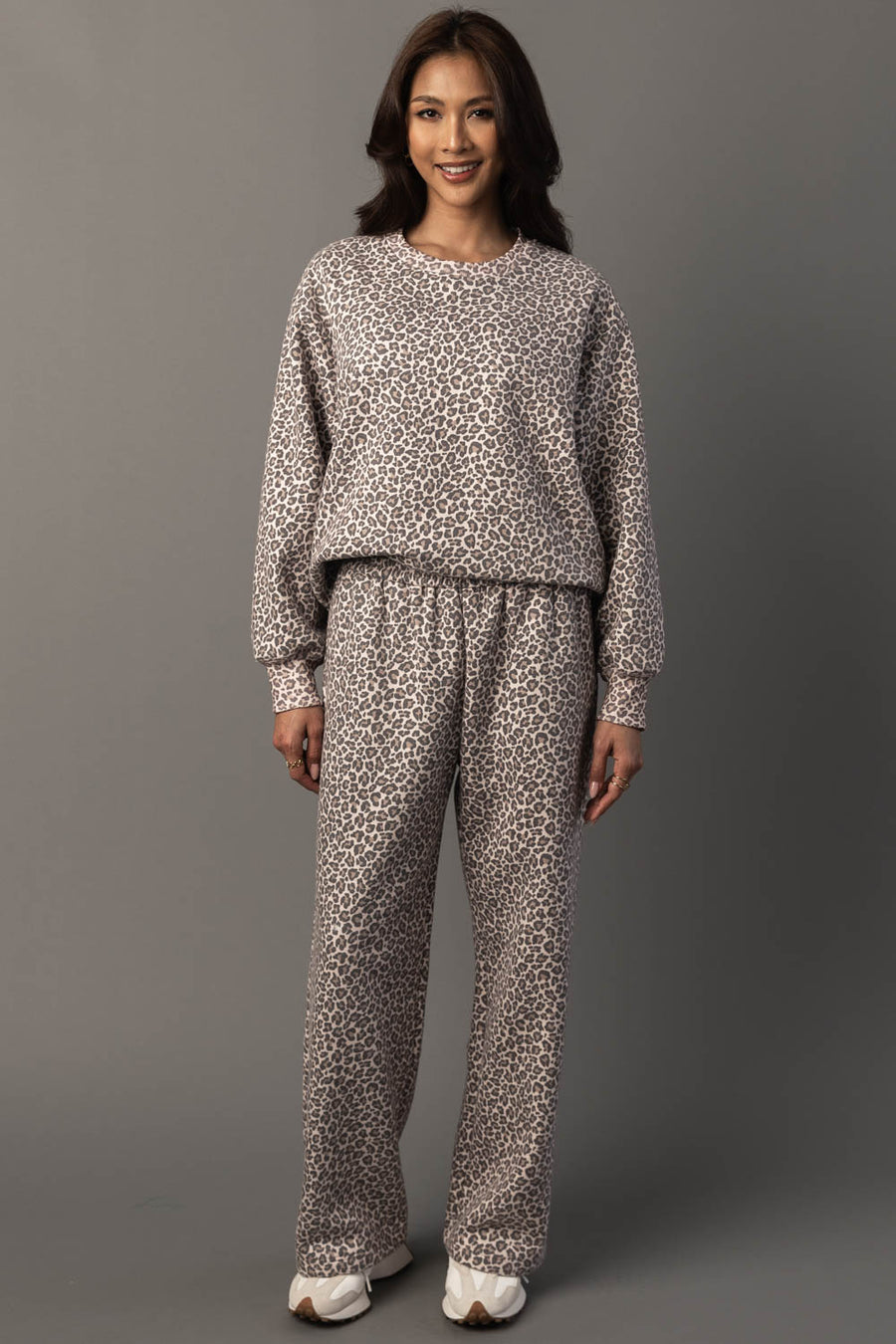 A woman stands smiling against a plain gray background, wearing the Panache Apparel Collection Cream & Brown Leopard Sweatshirt paired with matching pants and white sneakers.