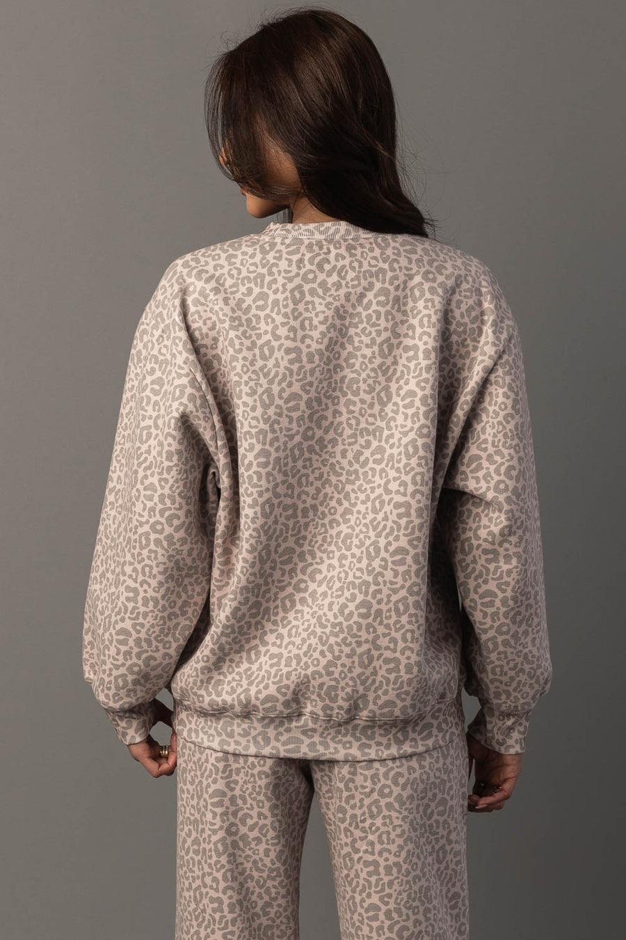 A person with long brown hair is seen from behind against a plain gray background, wearing the Panache Apparel Collection’s Blush & Light Brown Leopard Sweatshirt with matching pants.
