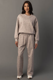A woman with long dark hair stands against a gray background, wearing the Panache Apparel Collection Blush & Light Brown Leopard Sweatshirt and matching pants set, paired with white sneakers. She has a neutral expression and her arms are relaxed by her sides.