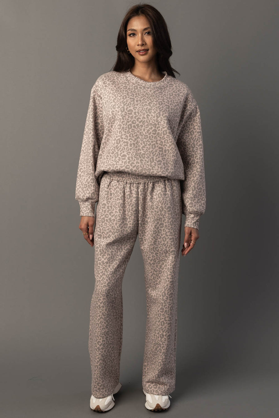 A woman with long dark hair stands against a gray background, wearing the Panache Apparel Collection Blush & Light Brown Leopard Sweatshirt and matching pants set, paired with white sneakers. She has a neutral expression and her arms are relaxed by her sides.