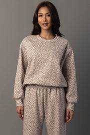 A woman stands against a gray background, wearing the Panache Apparel Collection Blush & Light Brown Leopard Sweatshirt with matching sweatpants, smiling slightly in a relaxed pose.