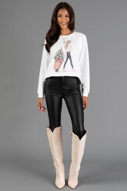 A woman wears a white long-sleeve Girl with Christmas Tree in Basket Sweatshirt from the Panache Apparel Collection, featuring an illustration of a girl pulling a basket with a decorated tree, paired with black pants and tall beige boots.
