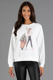 A woman wears a white Girl with Christmas Tree in Basket Sweatshirt featuring an illustration of a girl pulling a Christmas tree in a basket.