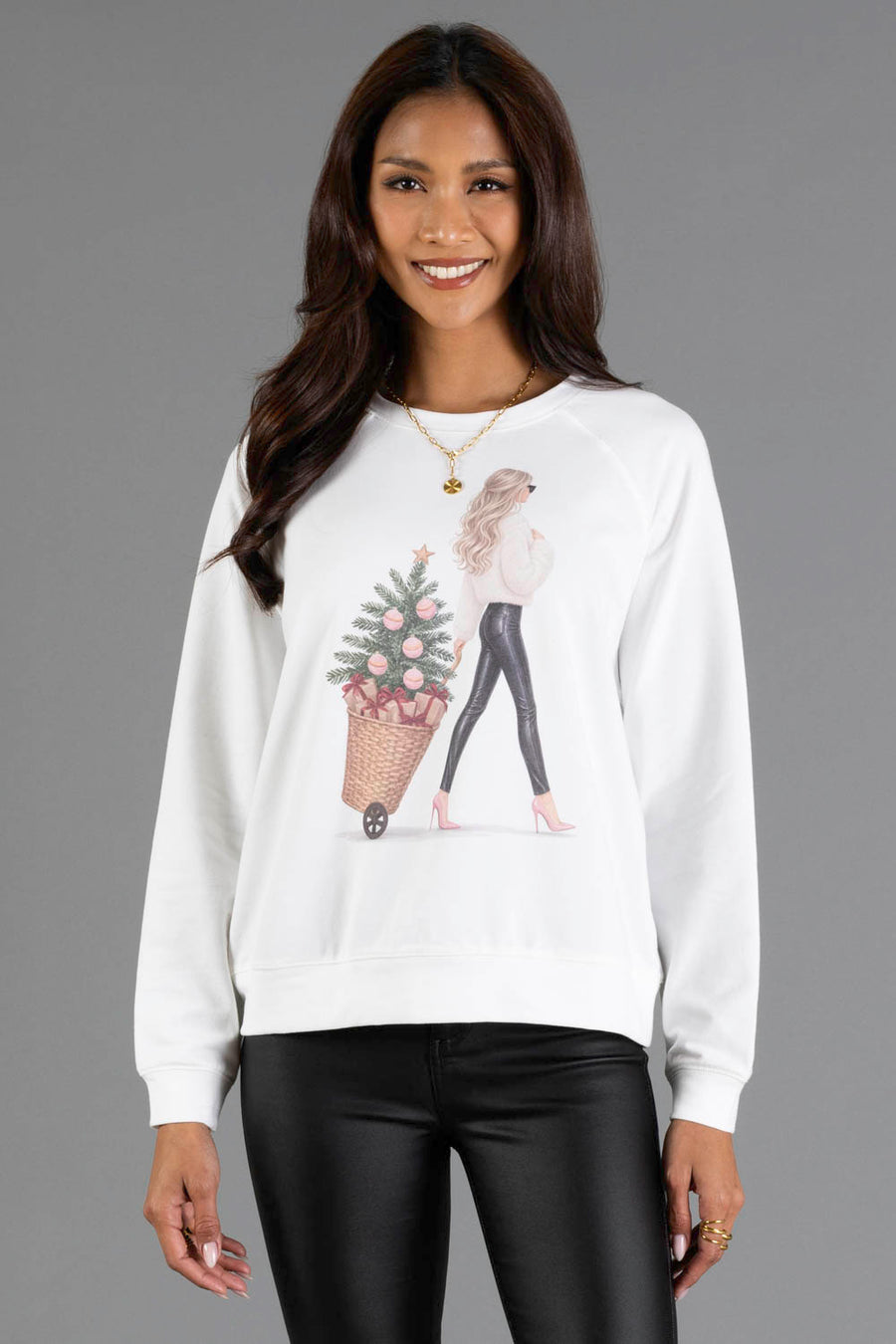 A woman wears a white Girl with Christmas Tree in Basket Sweatshirt featuring an illustration of a girl pulling a Christmas tree in a basket.
