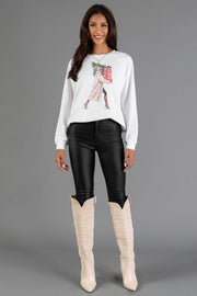 A woman wears a white Girl Carrying Presents & Tree Sweatshirt from the Panache Apparel Collection featuring a graphic of a woman carrying a Christmas tree and presents, paired with black pants and tall beige boots.
