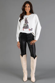 A woman wears a white Girl Walking Dog with Christmas Wreath Sweatshirt from the Panache Apparel Collection, featuring an illustration of a person walking a small dog while carrying a holiday wreath, paired with black pants and knee-high boots.