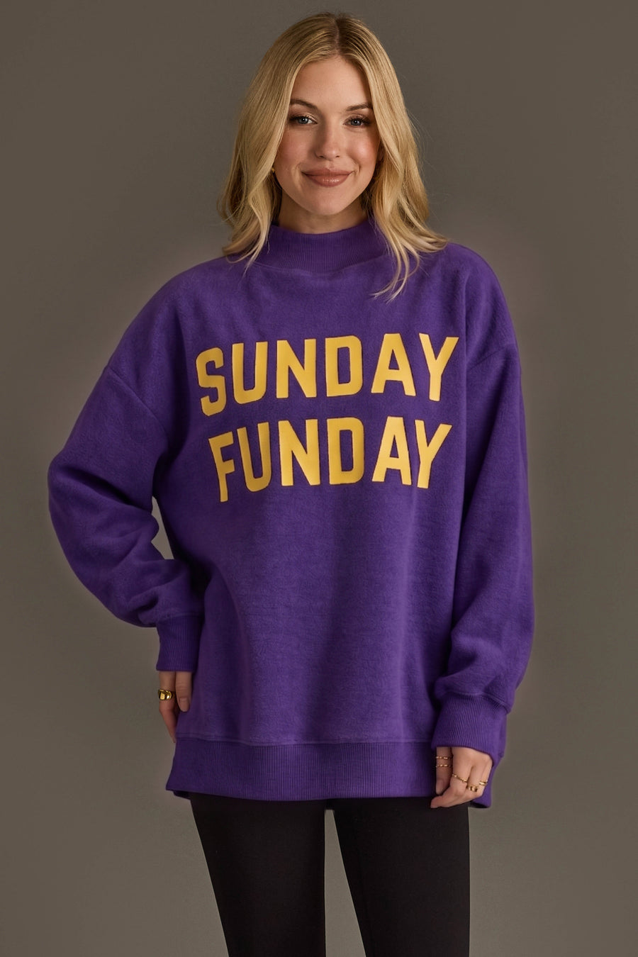 A smiling woman models the Panache Apparel Collection’s Purple Game Day & Sunday Funday Reversible Sweatshirt, featuring bold yellow text, styled with black pants against a plain grey background—ideal for ultimate game day comfort.