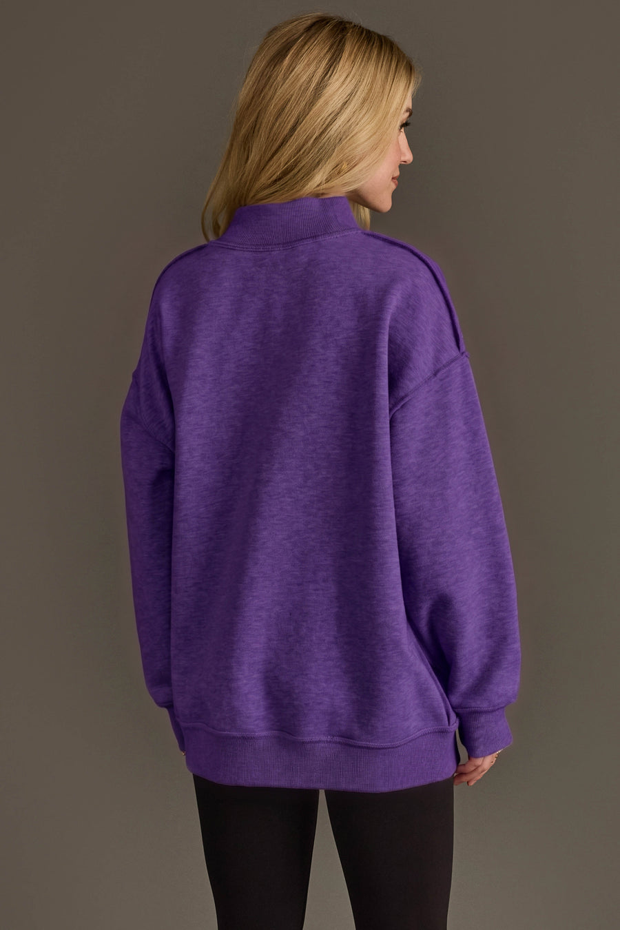A woman with long blonde hair stands with her back to the camera against a plain background, wearing the Panache Apparel Collection’s Purple Game Day & Sunday Funday Reversible Sweatshirt and black leggings.